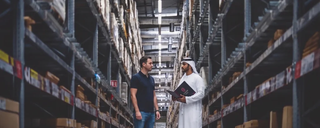 An Emirati and Western businessman meet in a busy Dubai warehouse, with a Crimson Legal booth prominent