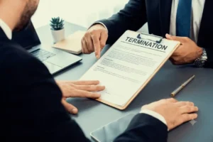 Two businessmen sit across a desk. One hands a clipboard displaying a formal TERMINATION document to the other.