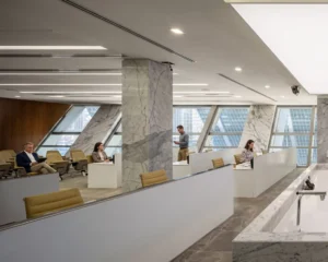 Modern open-plan office with marble column and city view windows, showing people working at desks and sitting in lounge chairs.