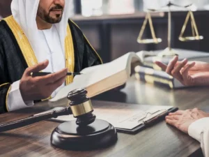 An Emirati judge in traditional attire sits in a courtroom, holding a pen and an open book, with a gavel in front of him and the scales of justice in the background